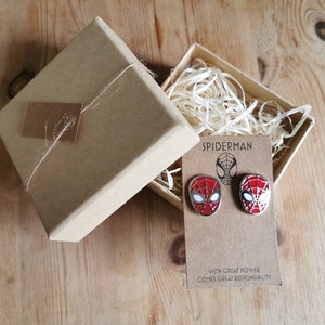 Op de afbeelding: Twee Spider-Man gezicht emaille pins op een kaart met de tekst "Spider-Man" en "With great power comes great responsibility." De pins zijn rood, wit en zwart, gepresenteerd in een kraft papieren geschenkdoos.
