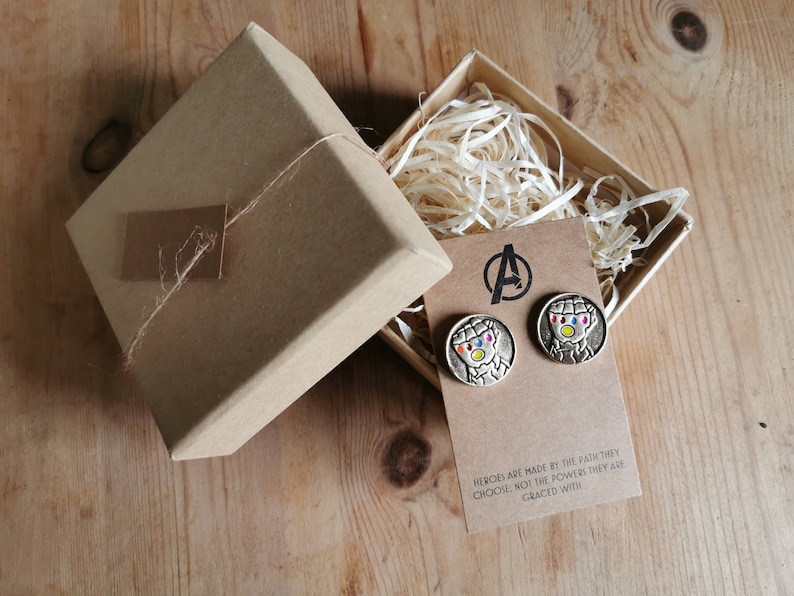 Op de afbeelding: Ronde, zilverkleurige oorstekers met een kleurrijk karakterontwerp, gepresenteerd op een bruine kaart met het Avengers-logo. De oorbellen worden getoond in een open geschenkdoos met decoratieve papieren vulling. Tekst op de kaart.
