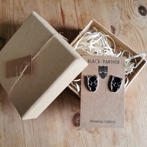 Op de afbeelding: Een bruine geschenkdoos met een paar Black Panther oorbellen. De oorbellen zijn zwart met een zilveren omtrek van het Black Panther masker. De tekst "Black Panther" en "Wakanda Forever" staat op de kaart.