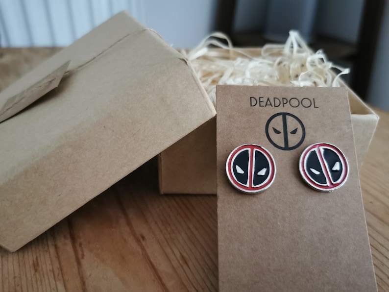 Op de afbeelding: Deadpool logo oorstekers met een zwart-wit ontwerp, gepresenteerd op een bruine kaart. De kaart zit in een kraft papieren geschenkdoos met versnipperd papier. Het woord "DEADPOOL" staat op de kaart.