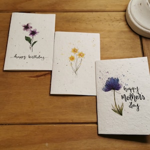 Może przedstawiać: Trzy ręcznie robione kartki z życzeniami z akwarelowymi ilustracjami kwiat&oacute;w. Pierwsza kartka zawiera napis "happy birthday" i przedstawia fioletowe kwiaty. Druga kartka przedstawia ż&oacute;łte kwiaty. Trzecia kartka zawiera napis "happy mother's day" i przedstawia niebieski kwiat.