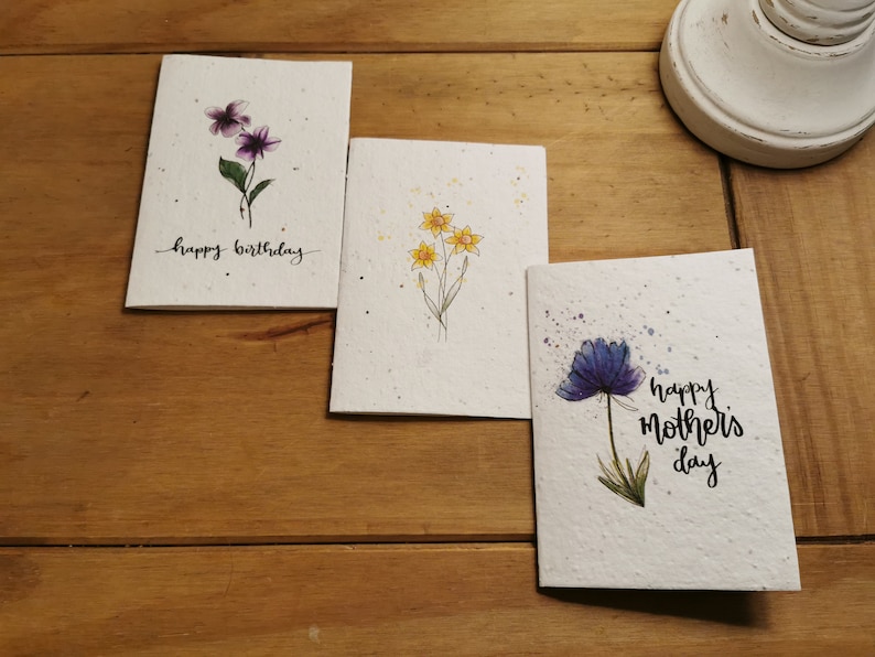 Może przedstawiać: Trzy ręcznie robione kartki z życzeniami z akwarelowymi ilustracjami kwiat&oacute;w. Pierwsza kartka zawiera napis "happy birthday" i przedstawia fioletowe kwiaty. Druga kartka przedstawia ż&oacute;łte kwiaty. Trzecia kartka zawiera napis "happy mother's day" i przedstawia niebieski kwiat.