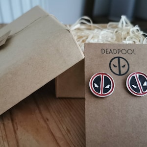 Puede incluir: Un par de pendientes de Deadpool de plata con esmalte rojo y negro. Los pendientes están empaquetados en una caja de cartón marrón con una etiqueta de papel marrón que dice "DEADPOOL".