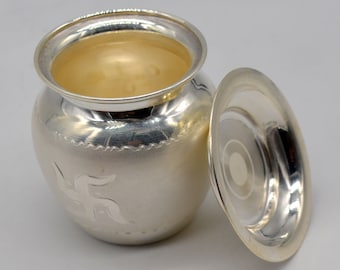 May include: A silver, round, decorative container with a lid. The container has a swastika symbol engraved on the front. The lid is slightly ajar.
