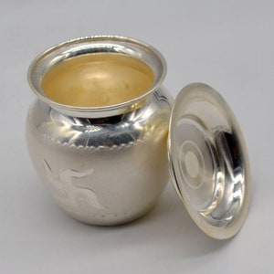 May include: A silver, round, decorative container with a lid. The container has a raised design of the Hindu symbol "Om" on the front. The lid is slightly ajar.