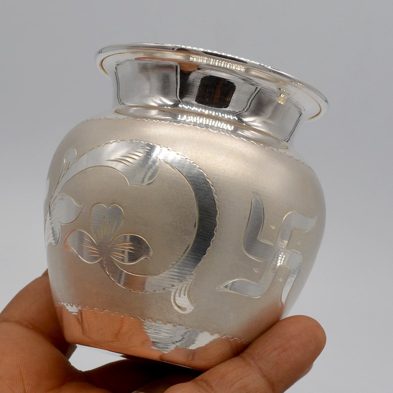 May include: A silver pot with intricate floral and swastika designs. The pot has a shiny, metallic finish and a rounded shape.