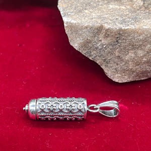 May include: A cylindrical silver pendant featuring an intricate embossed pattern of repeating scrollwork and rope-like borders. The pendant has a rounded end cap and a polished silver bail attached to one side, designed for suspension on a chain or cord.