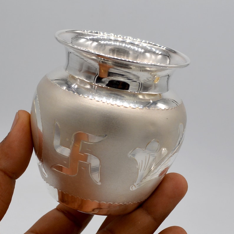May include: A silver pot with intricate designs, including a swastika symbol, on the surface. The pot is made of polished silver and has a rounded shape.