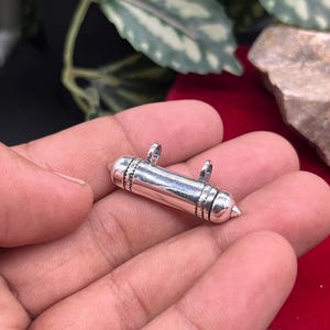 May include: A cylindrical silver pendant featuring a smooth, polished body with pointed, conical ends. The pendant includes two small, vertical loops attached to the top for suspension, and decorative etched detailing near the ends.