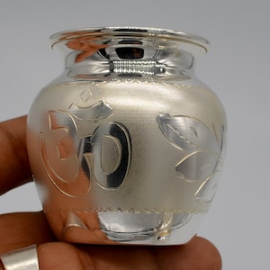 May include: A silver pot with an etched Om symbol and floral designs. The pot has a rounded body and a slightly flared rim.