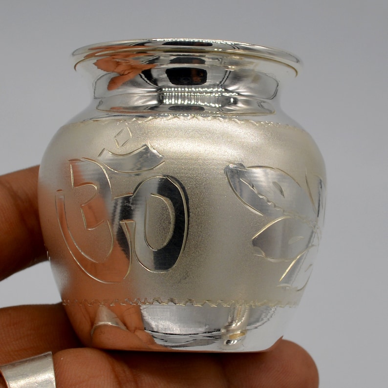 May include: A silver pot with an etched Om symbol and floral designs. The pot has a rounded body and a slightly flared rim.