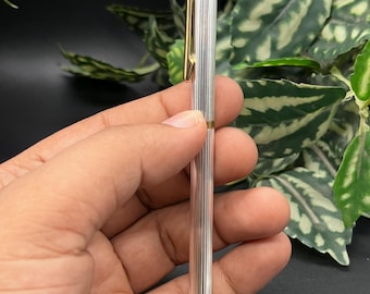Puede incluir: Un bolígrafo de tono plateado con una textura acanalada vertical a lo largo del cuerpo. El bolígrafo incluye un clip de bolsillo de color dorado en la parte superior y una punta cónica dorada a juego en la parte inferior, con acabado metálico.