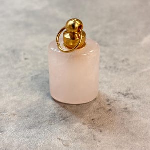 May include: A small, cylindrical rose quartz pendant with a gold-colored metal cap and loop. The pendant is a soft pink color and has a smooth, polished surface. The gold cap has a small, round top.