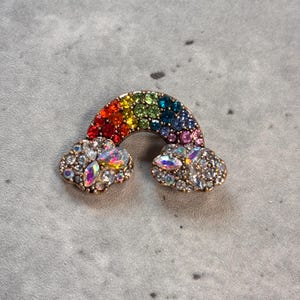 May include: A rainbow brooch with a gold-coloured metal frame. The rainbow is made of various coloured gemstones, and the clouds are adorned with iridescent crystals. This jewellery item is a fun accessory.