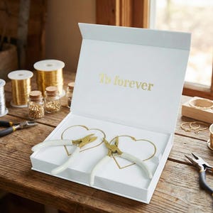 Könnte beinhalten: Eine weiße Schachtel mit dem goldenen Schriftzug "To forever". Im Inneren befinden sich zwei herzförmige Drahtdesigns und zwei Schmuckzangen mit weißen Griffen und goldfarbenen Köpfen. Golddrahtspulen und kleine Gläser mit Perlen sind ebenfalls zu sehen.