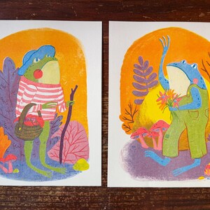 May include: Two rectangular art prints featuring anthropomorphic frogs. The left print shows a green frog in a blue hat and striped shirt holding a basket and walking stick. The right print shows a blue frog in green overalls holding flowers.