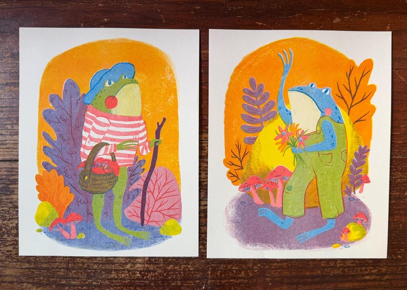 May include: Two rectangular art prints featuring anthropomorphic frogs. The left print shows a green frog in a blue hat and striped shirt holding a basket and walking stick. The right print shows a blue frog in green overalls holding flowers.