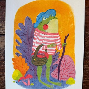 May include: A colorful illustration of a green frog wearing a blue hat and a pink and white striped sweater. The frog holds a brown woven basket filled with red mushrooms and a dark brown walking stick, standing before orange foliage.