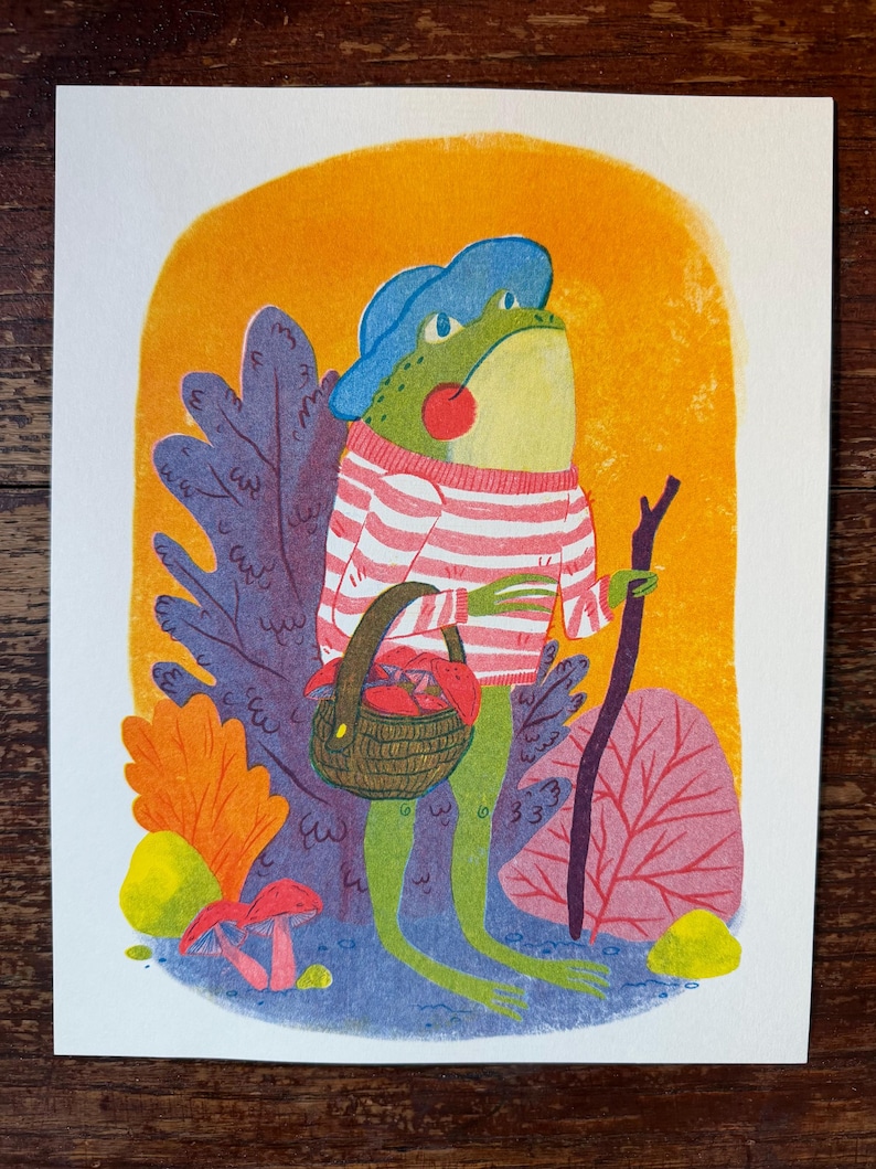May include: A colorful illustration of a green frog wearing a blue hat and a pink and white striped sweater. The frog holds a brown woven basket filled with red mushrooms and a dark brown walking stick, standing before orange foliage.