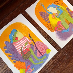 May include: Two rectangular art prints featuring colorful illustrations of frogs. One frog wears a striped sweater and carries a basket of mushrooms and a walking stick. The other frog wears green overalls and holds a bouquet of flowers near pink mushrooms.