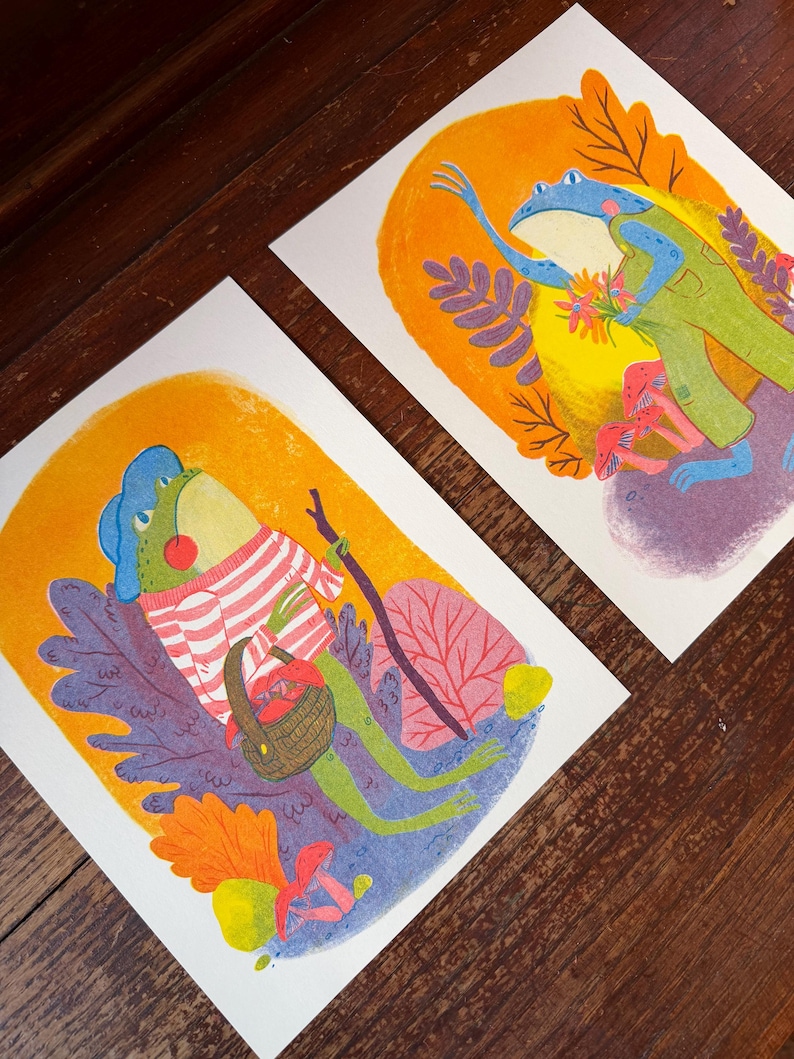May include: Two rectangular art prints featuring colorful illustrations of frogs. One frog wears a striped sweater and carries a basket of mushrooms and a walking stick. The other frog wears green overalls and holds a bouquet of flowers near pink mushrooms.
