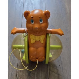 May include: A vintage orange plastic bear toy with blue eyes and a yellow bow tie. The bear is centered between two clear plastic wheels with lime green accents. A thin yellow string is attached to the bottom of the bear for pulling.