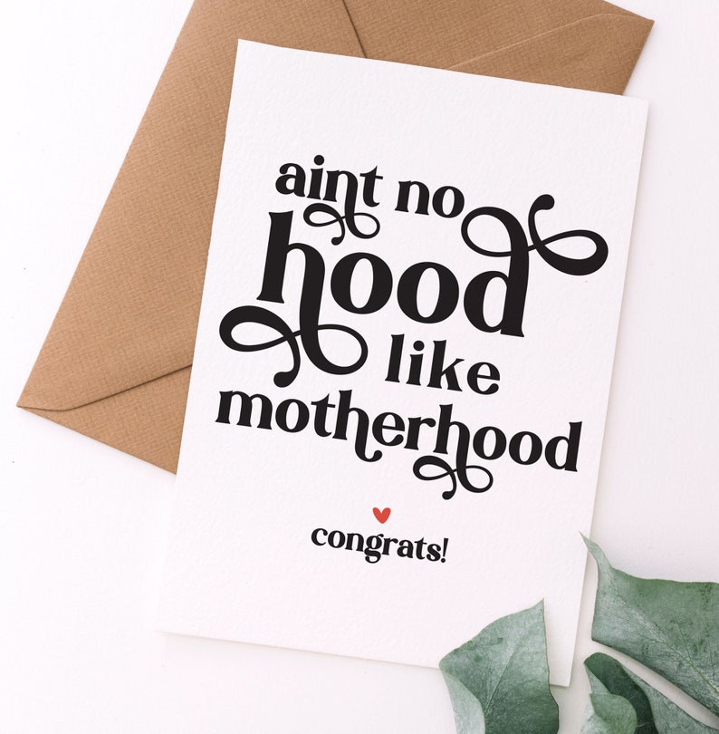 May include: A white greeting card with the text "aint no hood like motherhood" in a bold, black script. The word "congrats!" is printed below in a smaller font, with a red heart. A brown envelope is visible.