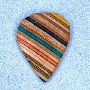 May include: A guitar pick made from recycled skateboard wood. The pick has a teardrop shape and is multi-colored with stripes of brown, yellow, blue, and green.