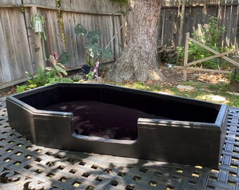 May include: A black coffin-shaped pet bed with a plush, dark purple interior. The bed is made of wood and sits on a decorative metal table outdoors, with a natural background of trees and a wooden fence.