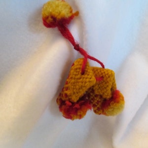 May include: A handmade, miniature pair of yellow and red knitted mittens, connected by a red yarn string. The mittens have a textured appearance with red accents and a fluffy pom-pom at the top. The background is a soft, white fabric.