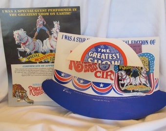 Può includere: Un cappello di carta blu e bianco con le parole "The Greatest Show" e "Ringling Bros. Circus" stampate su di esso. Il cappello è decorato con stelle e strisce rosse, bianche e blu. Il cappello è un'edizione speciale del bicentenario del circo Ringling Bros. e Barnum & Bailey.