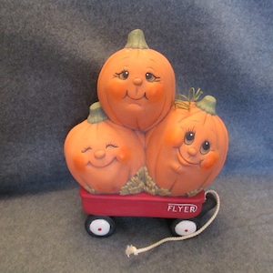 May include: Three orange pumpkins with painted faces are stacked in a red toy wagon with white wheels. The wagon has a white rope attached to it and the words "FLYER" printed on the side.