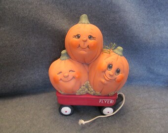 May include: Three orange pumpkins with painted faces are stacked in a red toy wagon with white wheels. The wagon has a white rope attached to it and the words "FLYER" printed on the side.