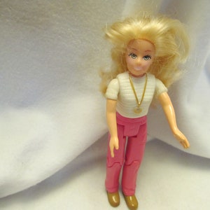 May include: A blonde doll with a white shirt, pink pants, and a gold necklace. The doll has a friendly expression and is wearing brown shoes.