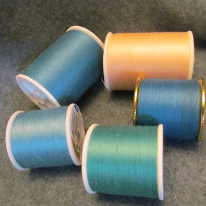 May include: A collection of colorful thread spools in shades of teal, peach, and green. The spools are arranged on a gray surface, with some having white or gold accents. Ideal for sewing and crafting projects.