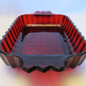 May include: A rectangular ruby red glass dish featuring a deep, pressed diamond pattern on the interior base and vertical fluted ridges along the exterior sides. The dish has scalloped edges and integrated handles on both ends of the vessel.