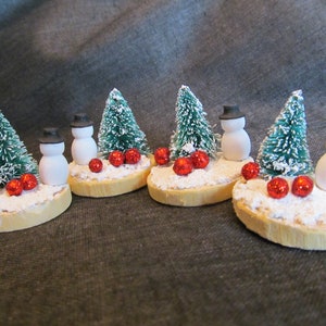 May include: Miniature Christmas scene featuring snowmen with black top hats, green bottle brush trees, and red glitter ball ornaments. The snowmen and trees are set on a wooden base with faux snow, creating a festive holiday decoration.