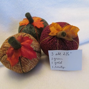 May include: Three fabric pumpkins, each approximately 2.5 inches in diameter, in green, gold, and rusty colors. The pumpkins have green stems and are adorned with orange and red fabric leaves. The pumpkins are handmade and suitable for fall decor.