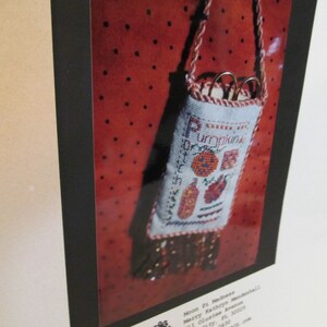 May include: A white needlepoint pouch with a red and green embroidered design of pumpkins and the text "Little Pumpkins". The pouch is hanging from a brown cord and has a beaded fringe.