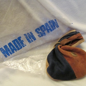 May include: A blue and brown suede pouch with a drawstring closure and a metal ring. The pouch is sitting on a clear plastic bag with the words "Made in Spain" printed in blue.