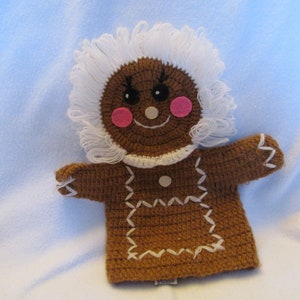 May include: A brown crocheted gingerbread person puppet with white yarn hair and pink felt cheeks. The puppet has a white crocheted collar and brown felt buttons.