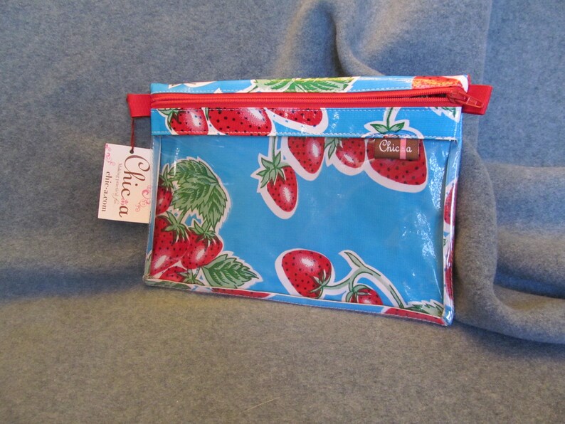 Puede incluir: Una bolsa azul con cremallera y un frente de vinilo transparente con un patr&oacute;n repetido de fresas rojas sobre un fondo blanco. La bolsa tiene una cremallera roja y una etiqueta blanca con la palabra "Chica" impresa en ella.