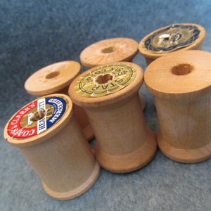 May include: Six wooden spools for thread, some with labels. The labels are in various colors and have text on them. The spools are all the same size and shape.