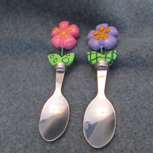 May include: Two decorative spoons with flower-shaped tops. One spoon has a pink flower with yellow accents, the other has a purple flower with yellow accents. Both spoons have green leaves and silver-colored spoon bowls.