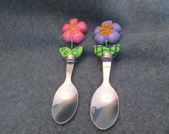 May include: Two decorative spoons with flower-shaped tops. One spoon has a pink flower with yellow accents, the other has a purple flower with yellow accents. Both spoons have green leaves and silver-colored spoon bowls.