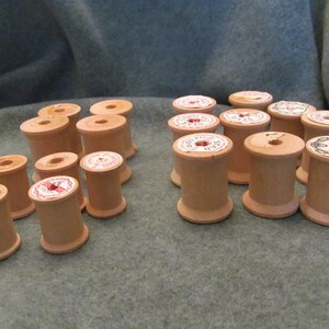 May include: A collection of wooden spools for thread, some with labels, in various sizes. The spools are arranged in two rows on a gray fabric surface.
