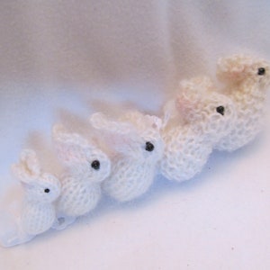 May include: Five white knitted bunny rabbits of varying sizes, arranged in a line from smallest to largest. The bunnies have black button eyes.