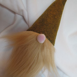 May include: A decorative gnome with a long, light brown beard and a pointed, olive green hat. The gnome has a small, pink nose. The beard is made of soft, fluffy material.