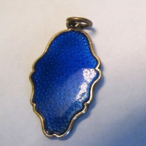May include: A small, blue enamel pendant with a gold-colored border and a textured surface. The pendant has a loop at the top for attaching to a chain or bracelet. The shape is irregular, with a rounded bottom and a more defined top.
