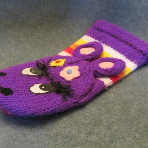 May include: Purple knitted puppet with a striped cuff, featuring a face with black eyes, a pink nose, and a yellow flower.